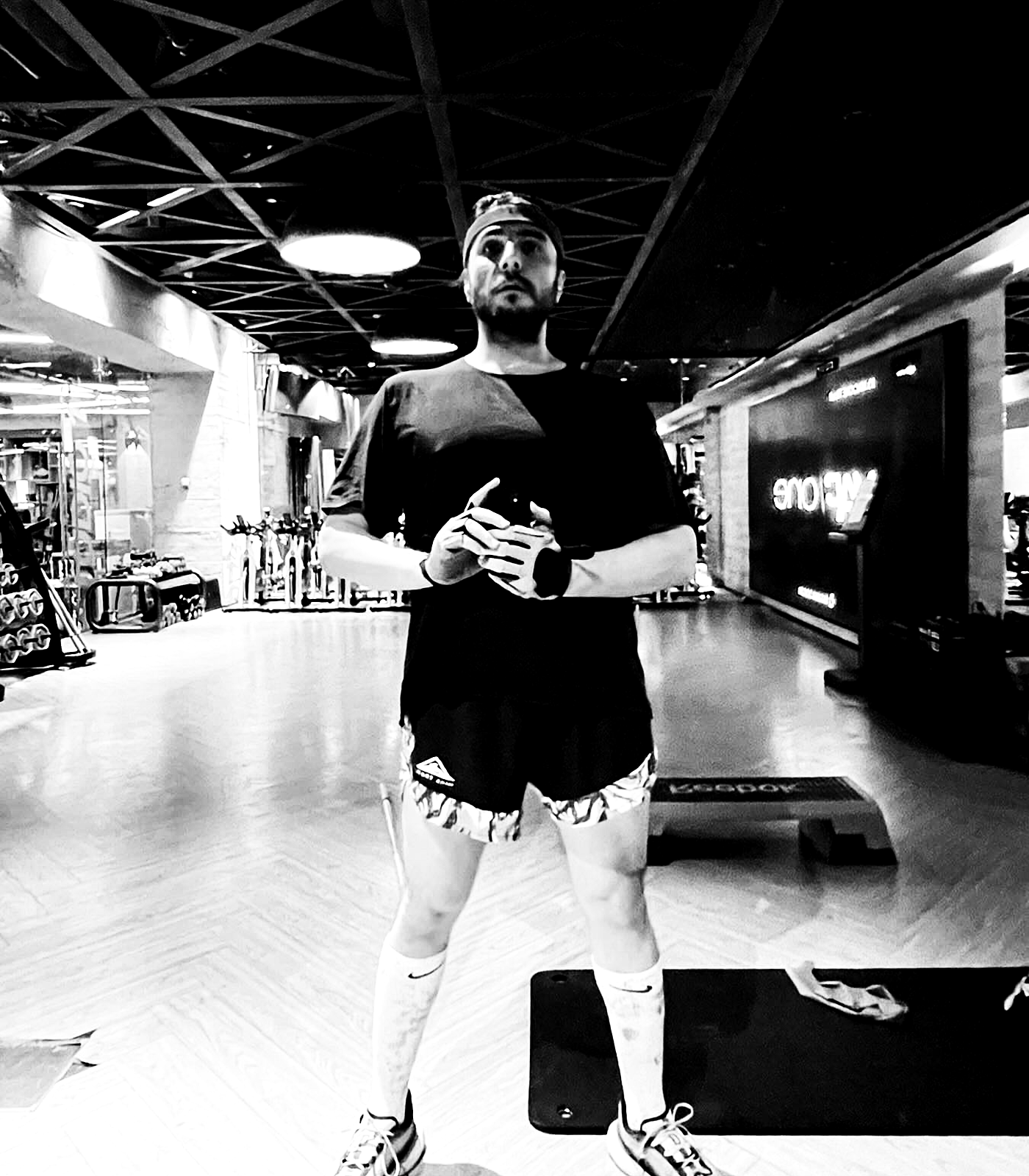 A full-body mirror portrait in a gym, centered under harsh ceiling lights with black shirt, shorts, and socks turned into hard graphic contrast.