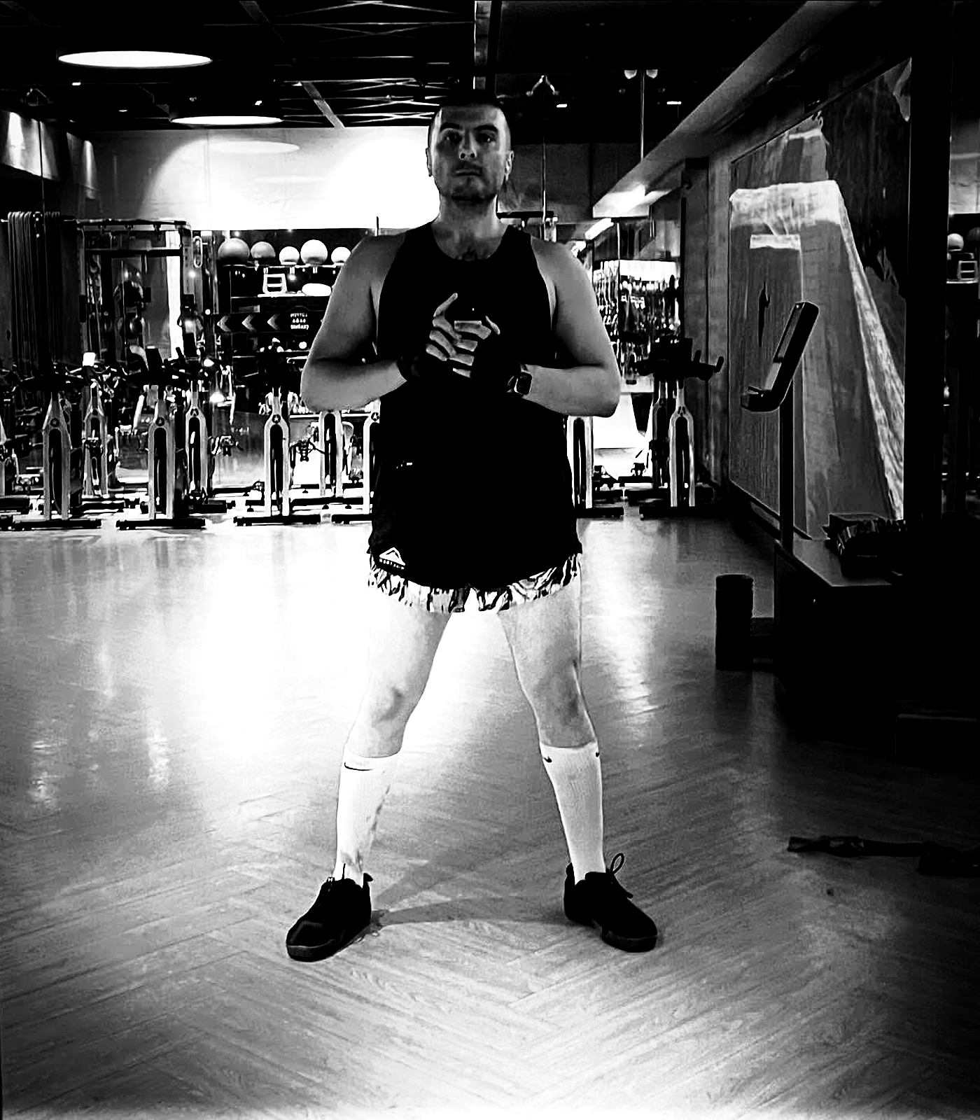 A full-body gym mirror image with feet planted wide and hands clasped at the center, rendered as a blunt high-contrast black-and-white plate.