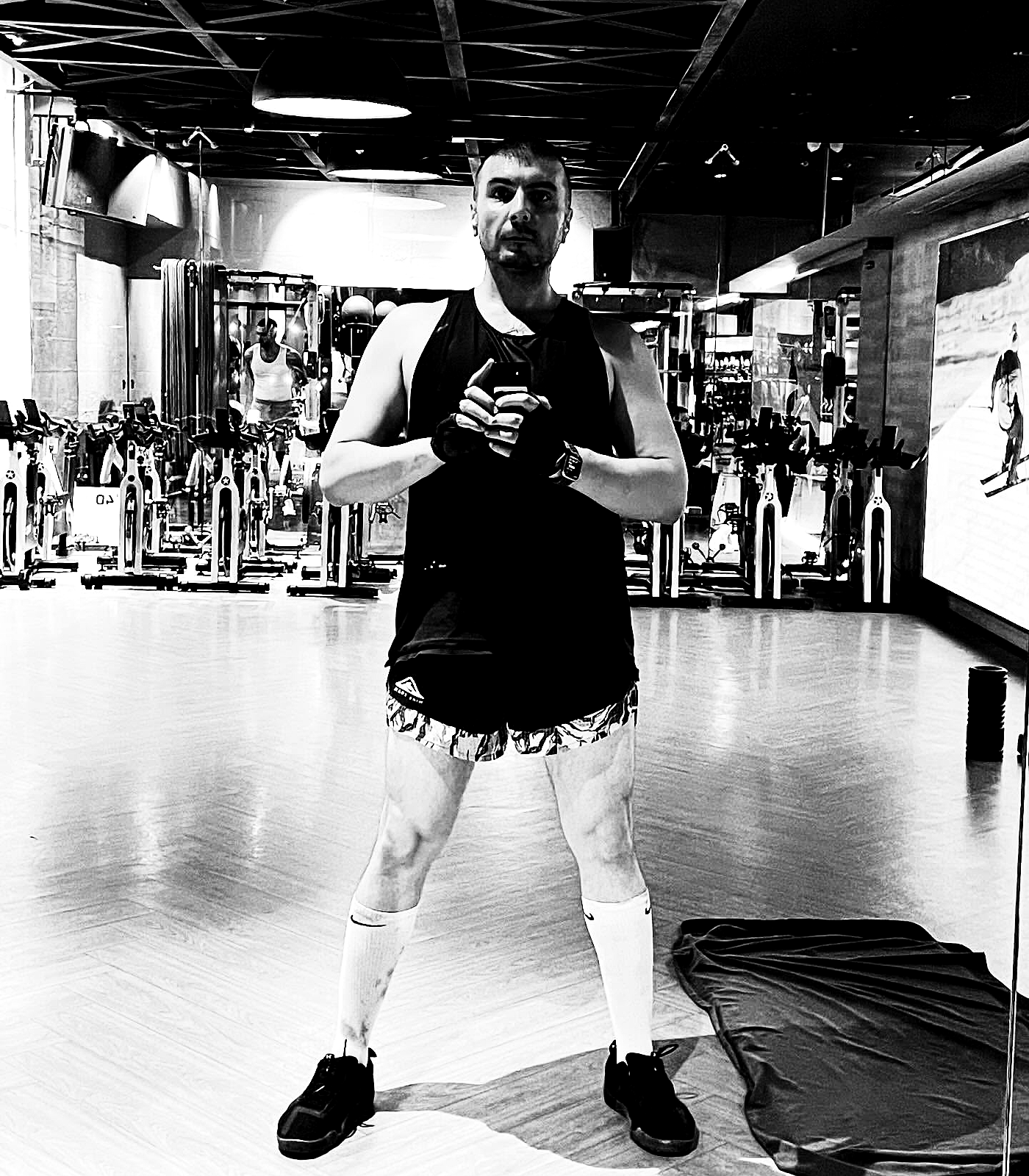A wide gym mirror portrait showing the subject in dark training clothes against an empty workout floor.