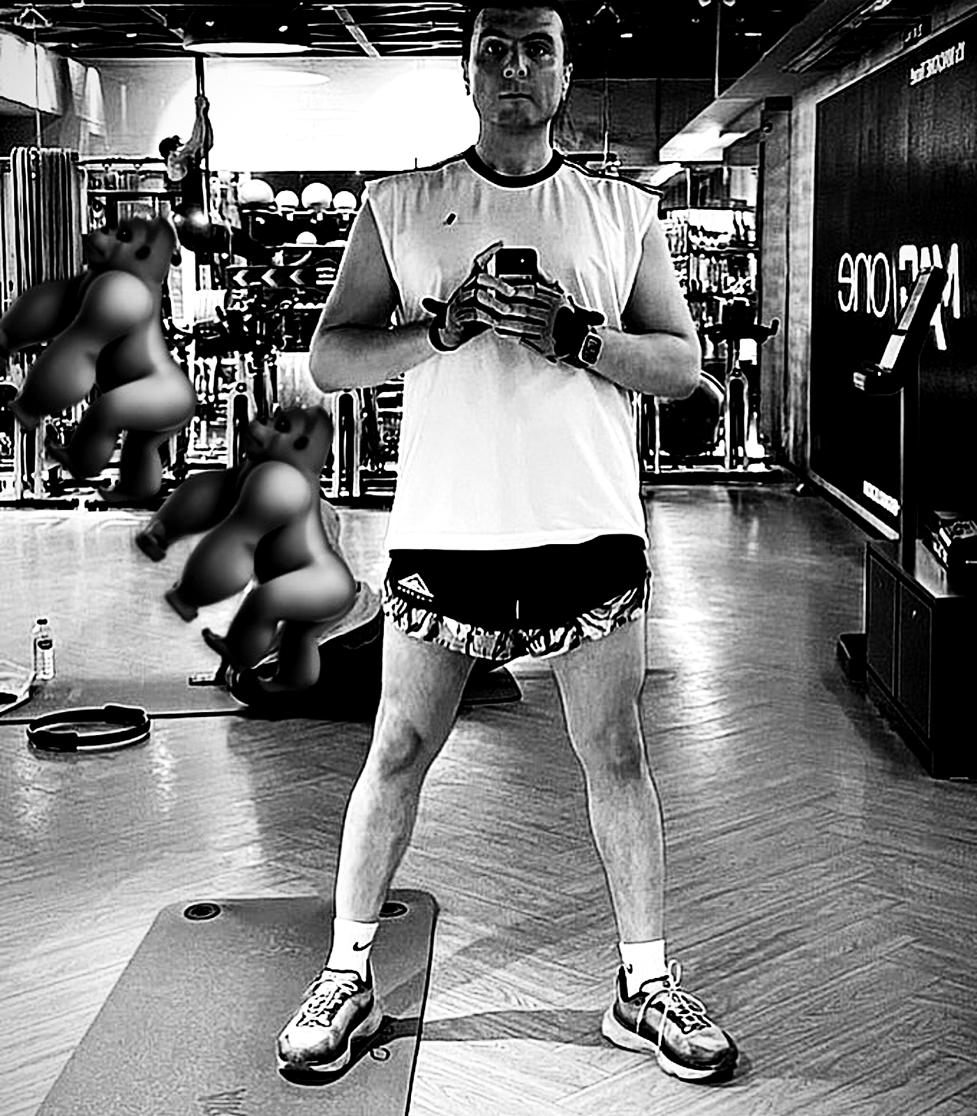 A full-body mirror portrait in a white shirt and dark shorts, with background figures softened and the gym floor flattened into glare.