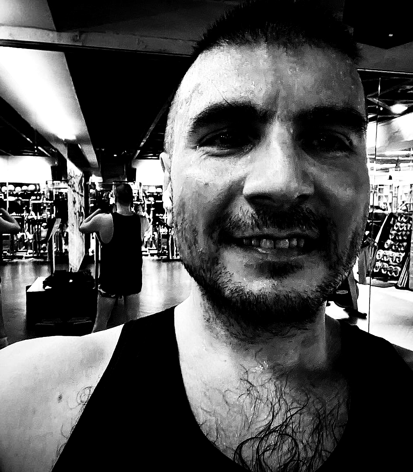 A close-up monochrome gym selfie with direct gaze, stubble, and fluorescent ceiling reflections pushed into harsh xerox contrast.