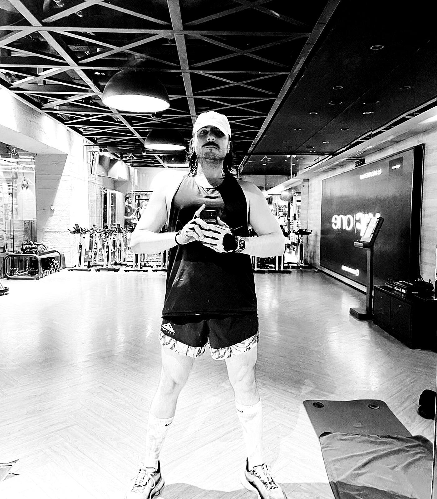 A wide full-body mirror portrait in a gym, framed by ceiling grid and equipment, turned into a stark repeated-performance image.